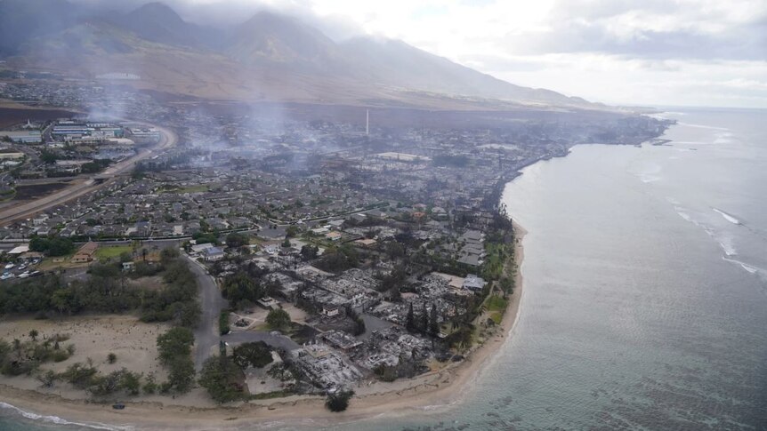 Aerial view from sea of Lahaina, a coastal town hit by deadly fires. Smoke billows from burnt out homes. Mountains behind town. 