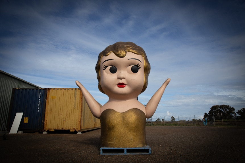 Giant kewpie doll torso with shipping containers and trees in background