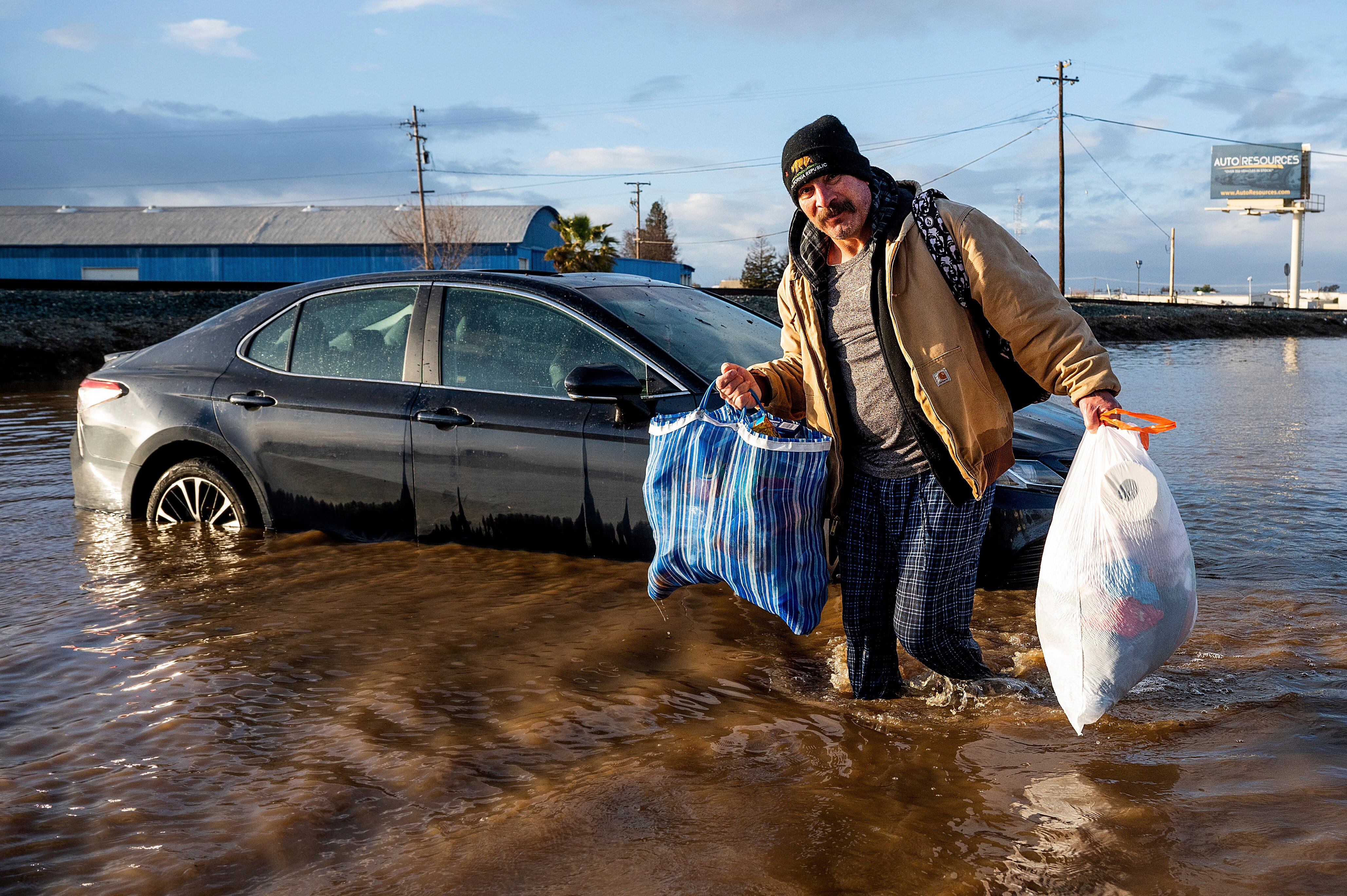 A man beside a car in flooded water carrying grocery bags