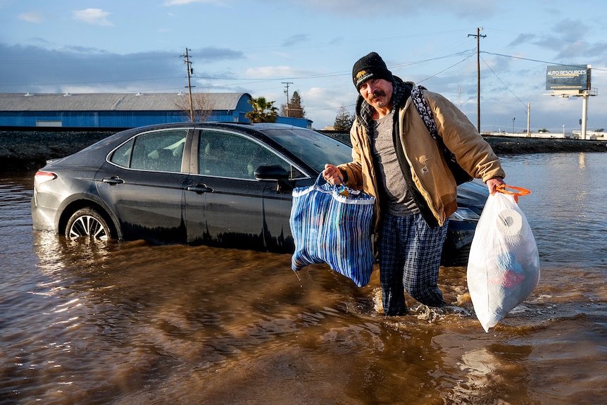 A man beside a car in flooded water carrying grocery bags