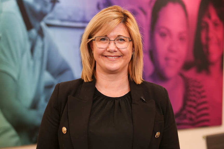 Claire Robbs is smiling at the camera in a portrait taken in front of a colourful mural featuring pictures of people.