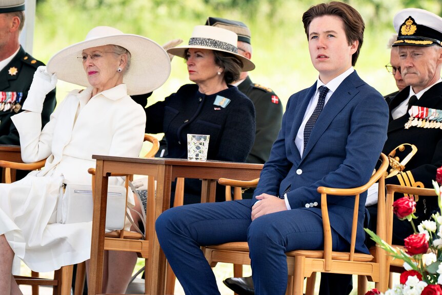 A woman in a white suit and a teenager in a blue suit sit in chairs in an outdoor audience