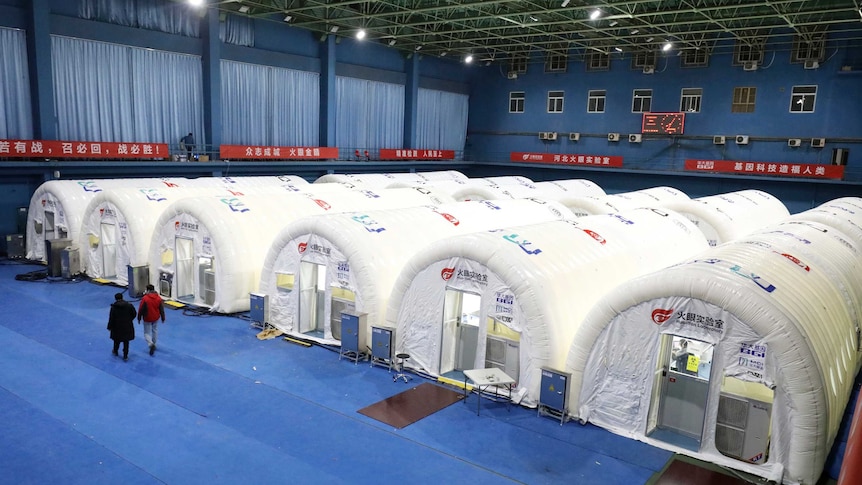 Workers walk past a temporary COVID-19 testing laboratory built on an indoor tennis court, there are six plastic huts.