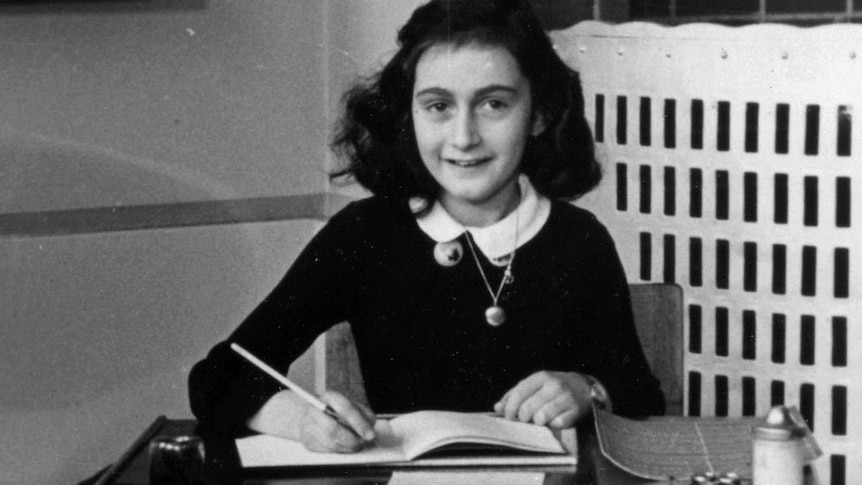 Anne Frank sits at a desk with a book open smiling at the camera.