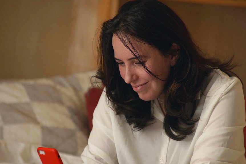A woman in a white jumper sits on a bed looking at a red phone