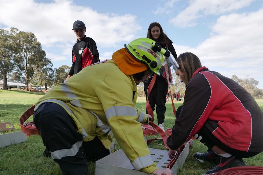 Three students, in red and black, assist a firefighter wearing yellow uniform to fold a red fire hose on a school oval.