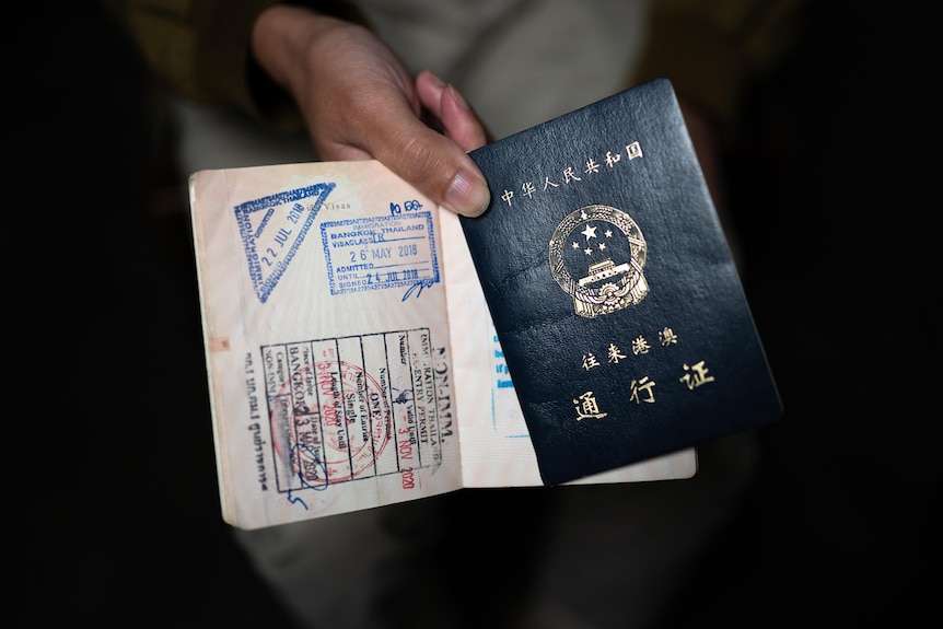 A person's hand holds a passport open at a page with travel stamps including Bangkok 