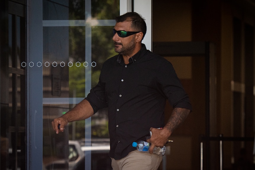A man wearing a navy button-up shirt and sunglasses walks out of court