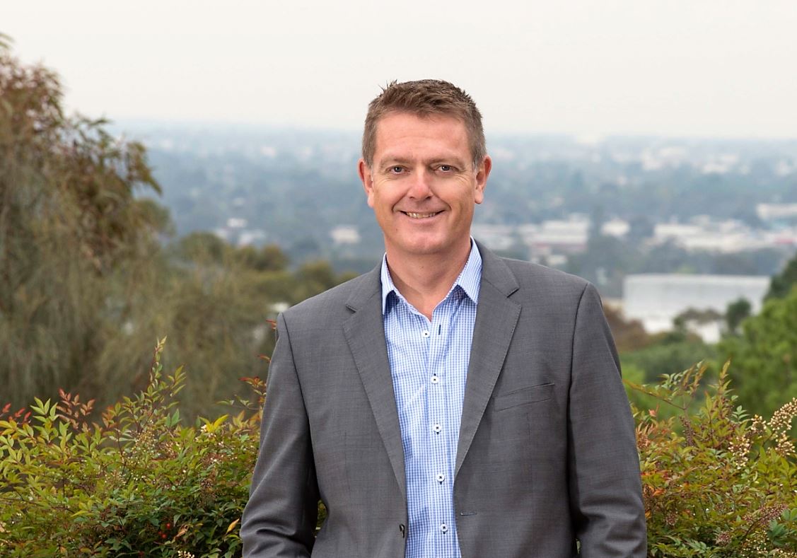 man in grey blazer and blue shirt smiles with bushes directly behind him and in distance city scape