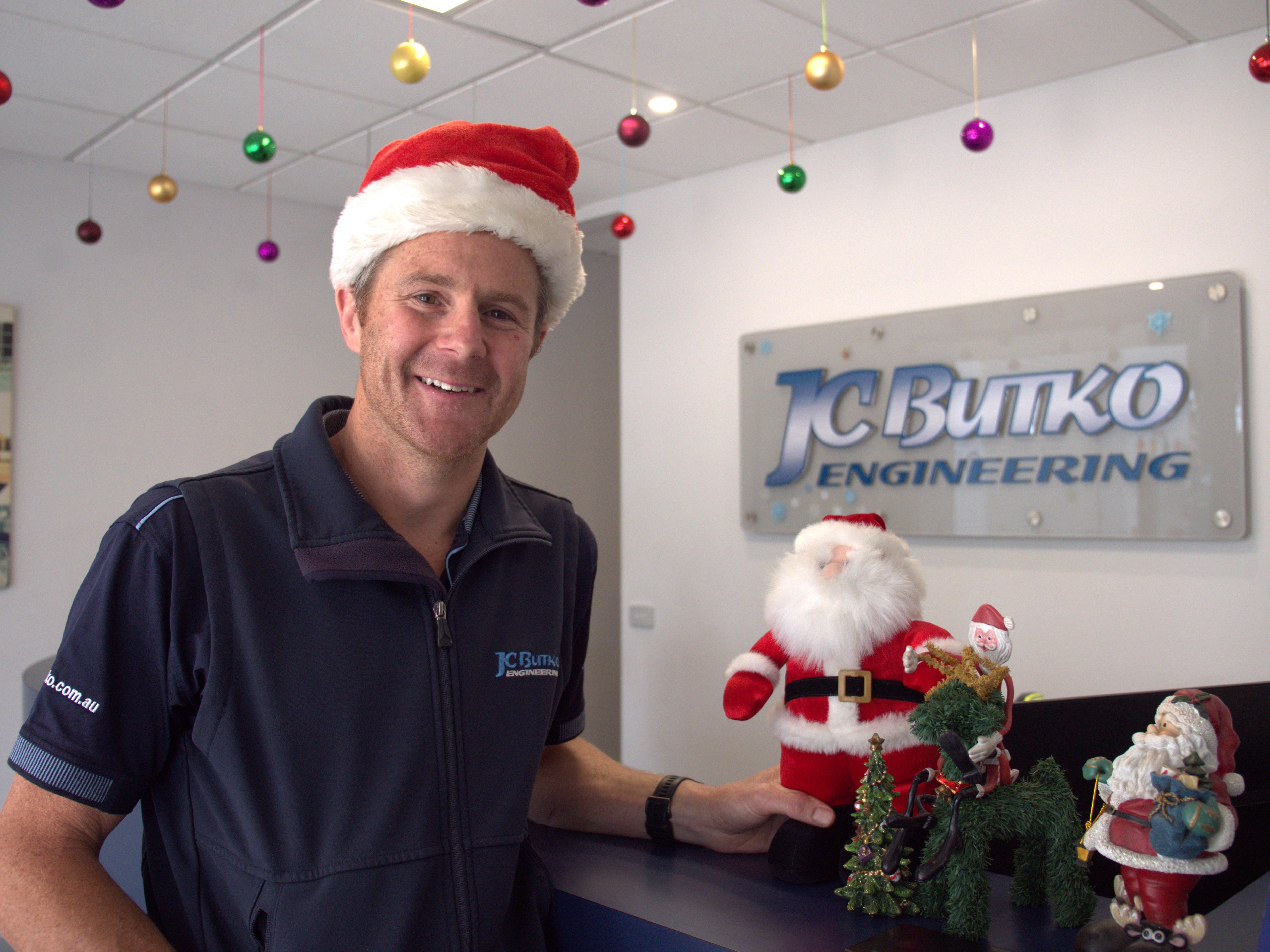 Paul  in a Santa hat and with a santa decoration at a desk smiles, his company logo JC Butco on wall.