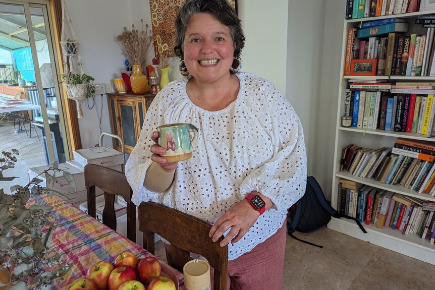 Sue Heward stands smiling at the dining table in her Monash home holding a tea cup.