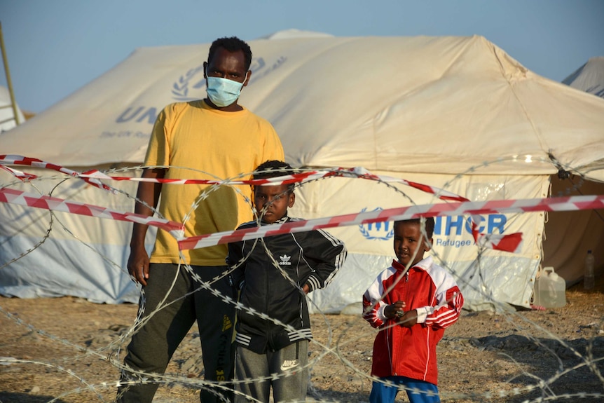 A man and two children stand behind razor wire at the new temporary refugee camp in Kara Tepe.