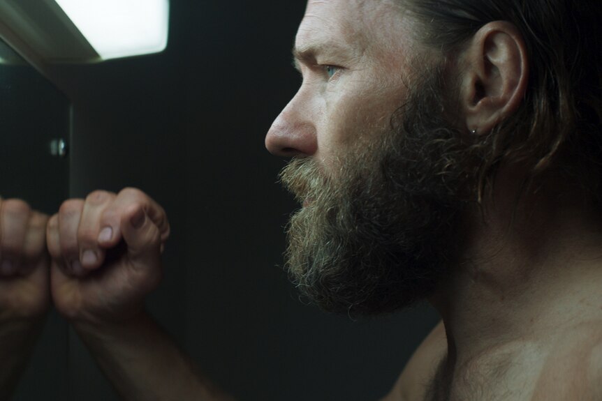 Profile of shirtless, middle-aged white man with rustic beard looking into mirror gloomily