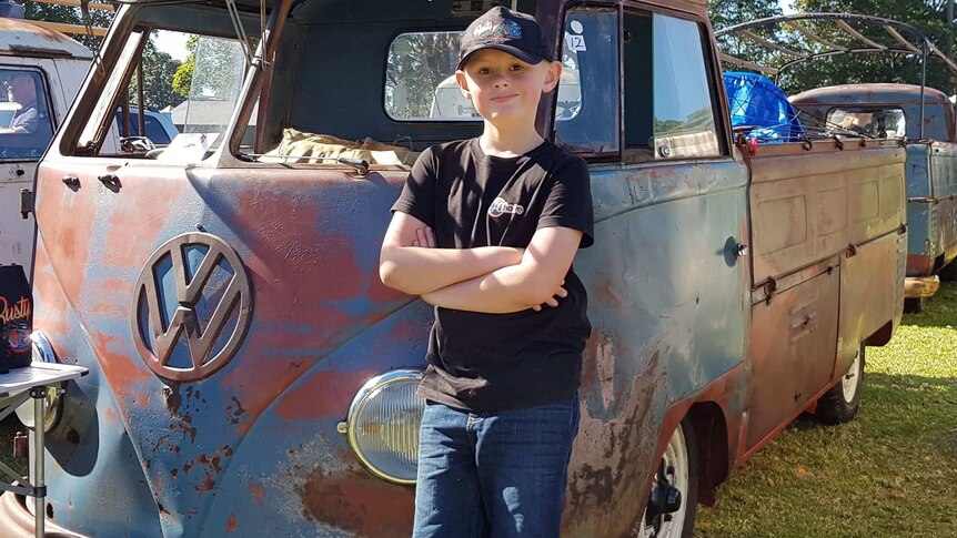Young boy in baseball cap stands beside rusty old Kombi