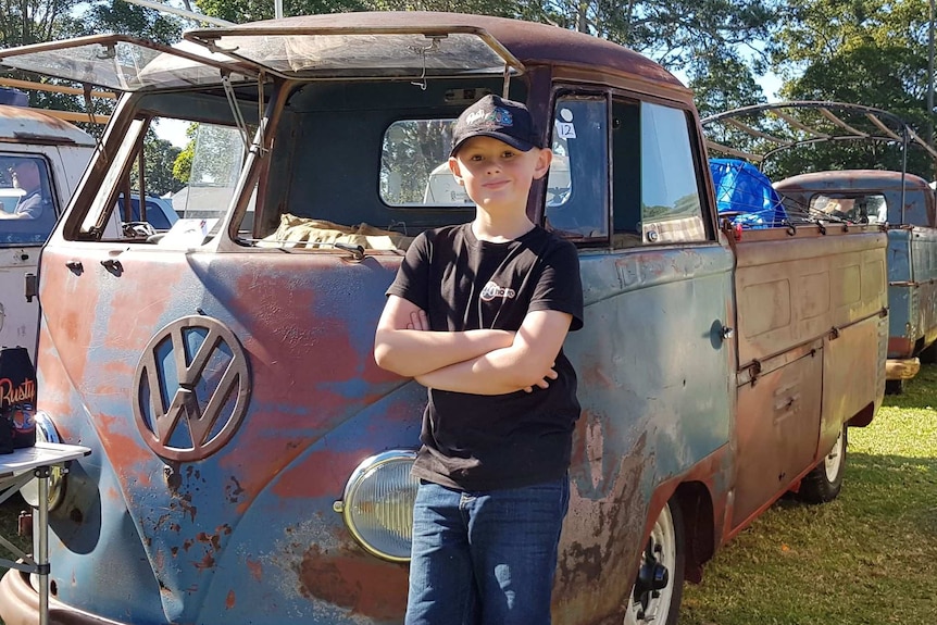 Young boy in baseball cap stands beside rusty old Kombi