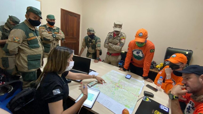 A dozen men in orange and green uniforms study a map on a conference table 