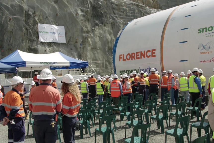 Dozens of people in high-vis and hard hats stand by a tunnelling machine several stories high. In front of it is a rockface.