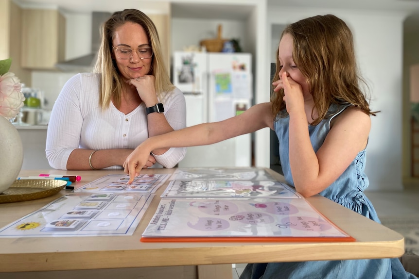 A women sits with her daughter at the kitchen table, looking at some organisational family charts. 