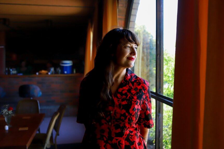 Woman wearing red and black dress stands by window looking out past curtain, with motel in background