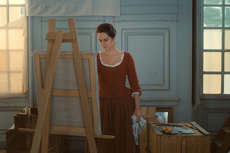 A woman wearing 18th century ochre dress stands in front of canvas and easel in an artist studio.