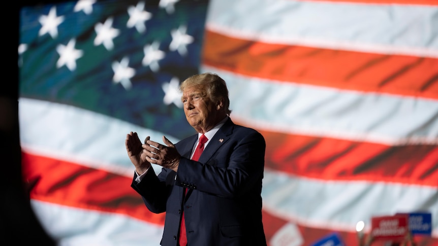 Donald Trump applauds in front of a US flag 