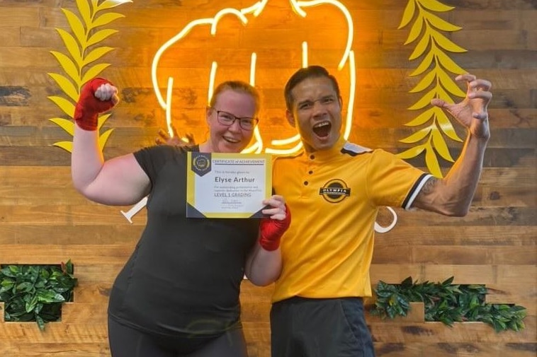 A woman with wraps on her hands stands next to a man in, she holds a certificate while they both flex their muscles