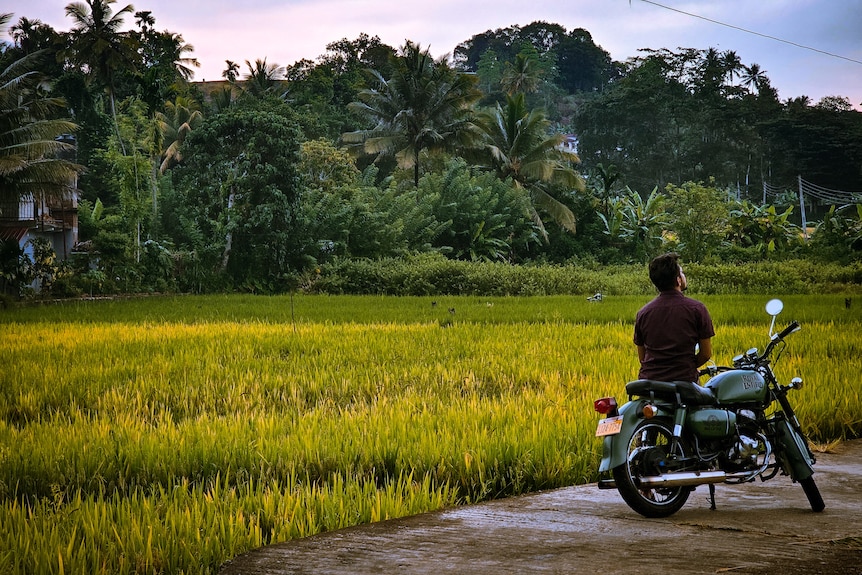 The back of a man resting on a motorbike in front of tropical-looking trees. 