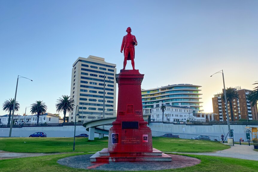 A large statue of Captain Cook covered in red paint, lit by dawn light in a St Kilda park.
