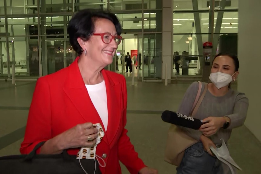 A woman wearing a red jacket walks past a reporter holding a microphone