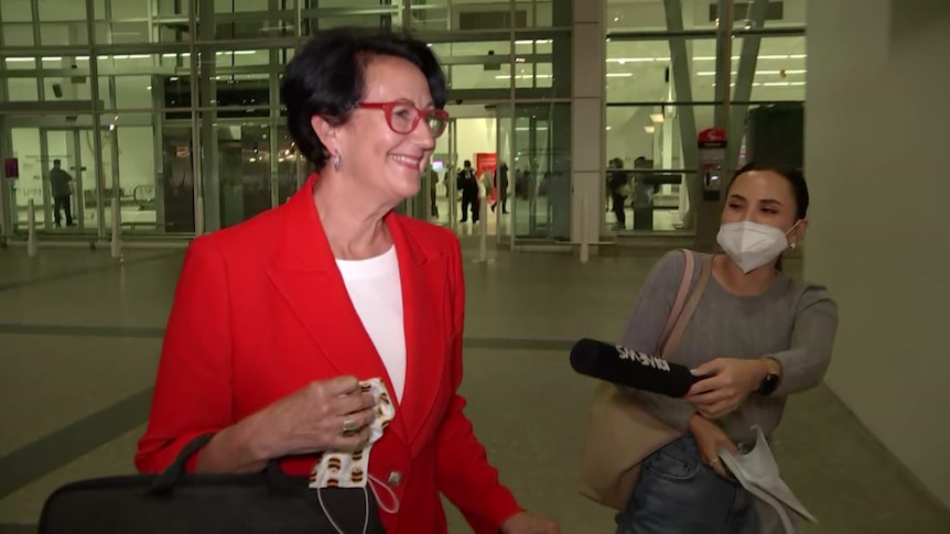 A woman wearing a red jacket walks past a reporter holding a microphone