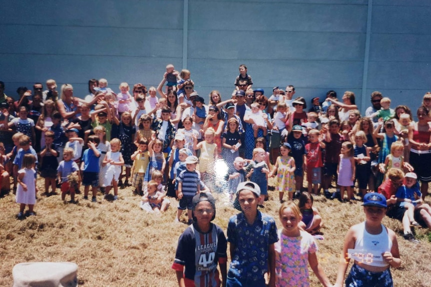 A large group of children with their families lined up against a grey concrete sound wall.
