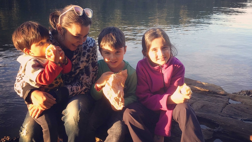 A mother sits by a river with her three children having a snack