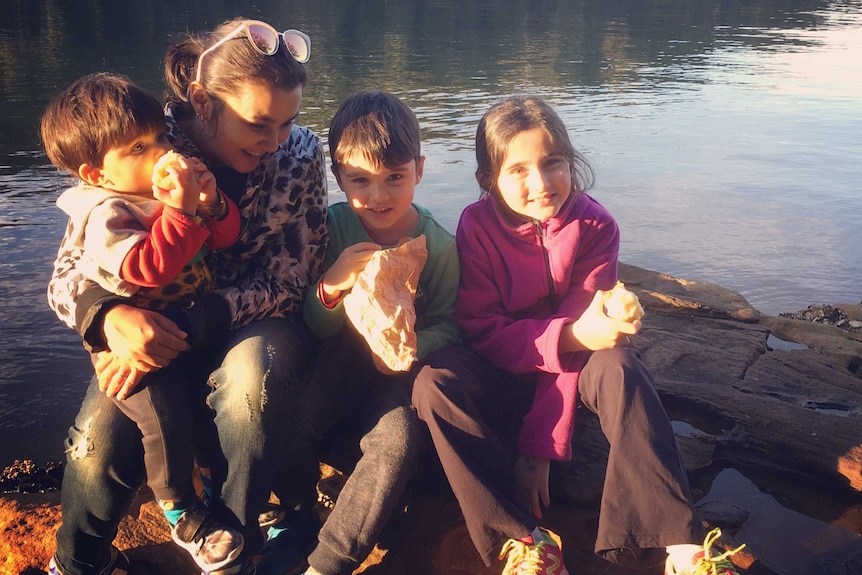 A mother sits by a river with her three children having a snack