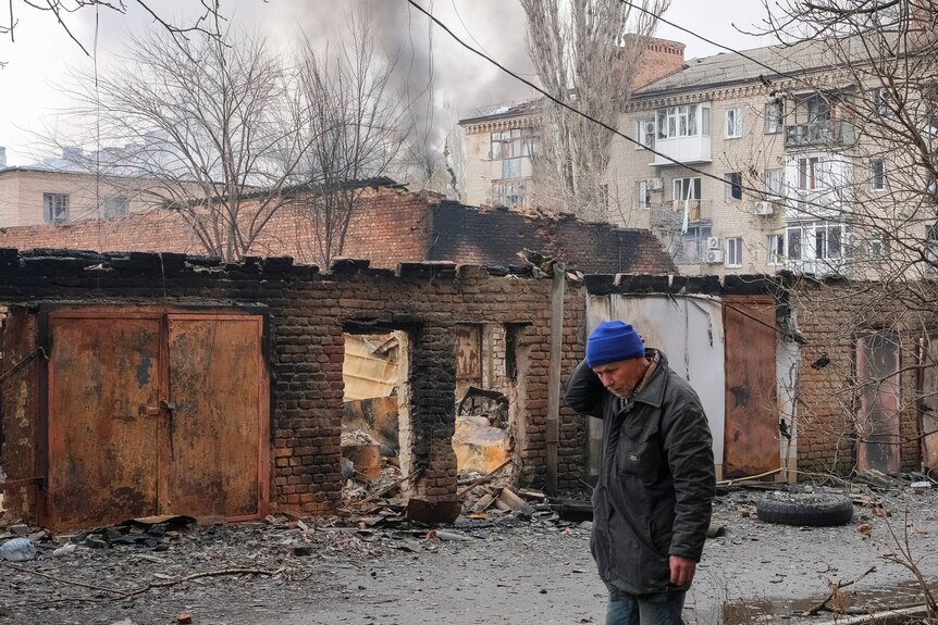 A man dressed in a coat and blue beanie stands in front of damaged buildings with smoke billowing in the distance.