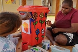 Two Aboriginal women sitting down outside painting a red bin