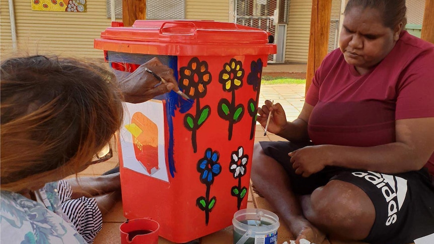 Two Aboriginal women sitting down outside painting a red bin