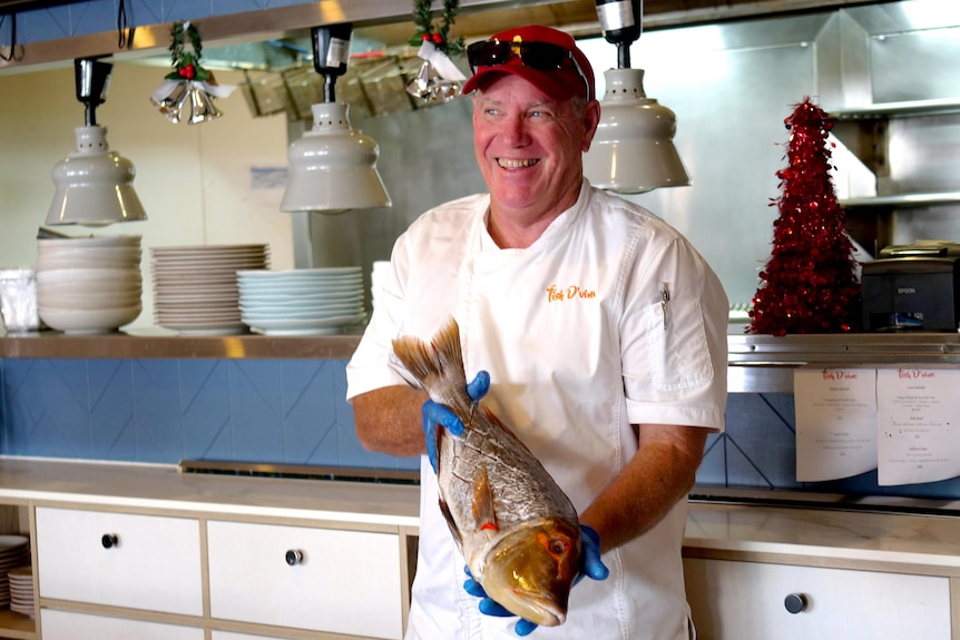 A man in chefs clothing stands holding fish, smiling and looking away from the camera in front of a restaurant kitchen