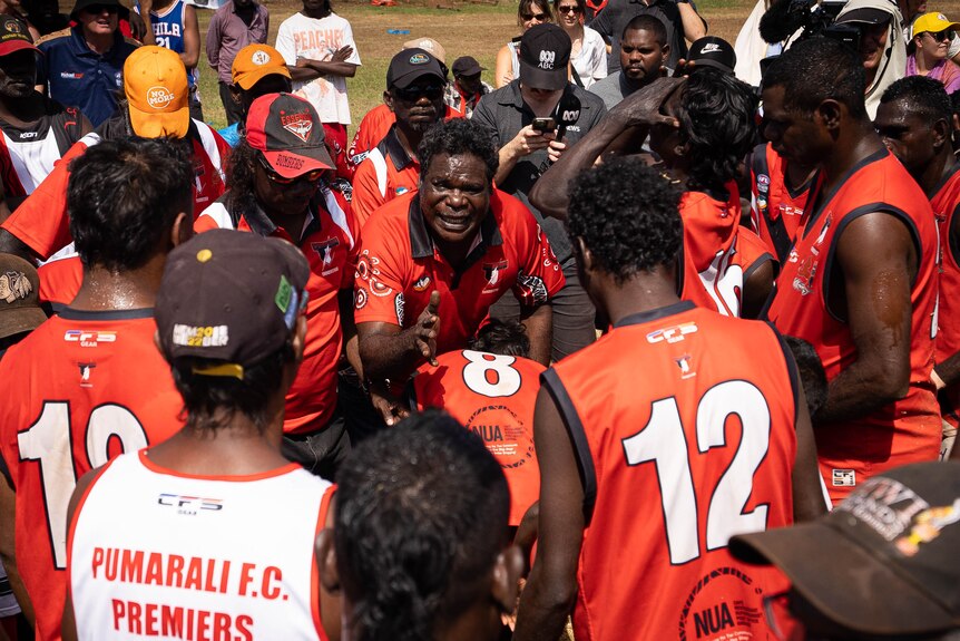 An Indigenous man talks sternly with a group of huddled footy players wearing identical jerseys