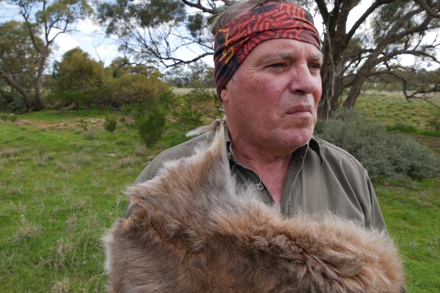 An Indigenous man wearing a bandanna stands outside, looking serious.