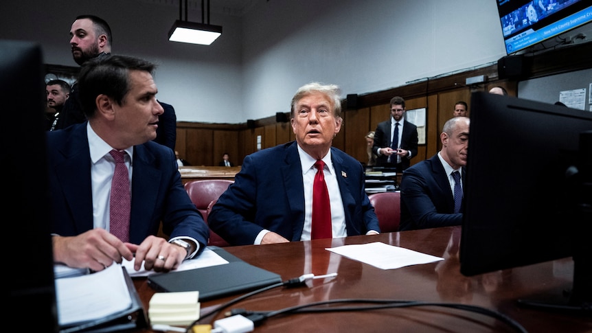 Donald Trump sits at a bench with men in suits either side of him. 