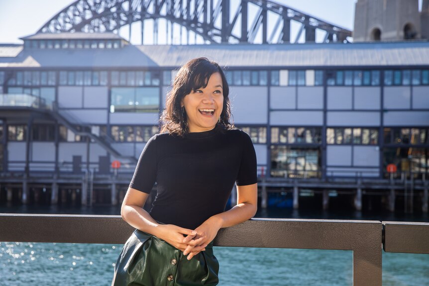 A smiling 30-something Asian Australian woman stands resting against a railing by the harbour, the Harbour Bridge behind her