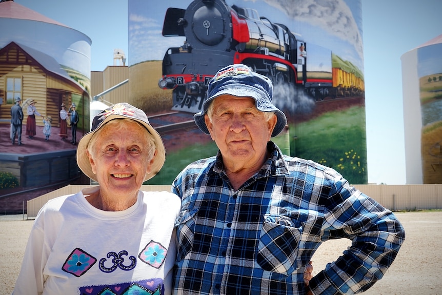 An elderly couple wearing bucket hats stand in front of wheat silos painted with art.
