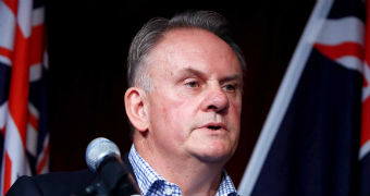 Mark Latham stands behind a microphone. There are two Australian flags behind him.