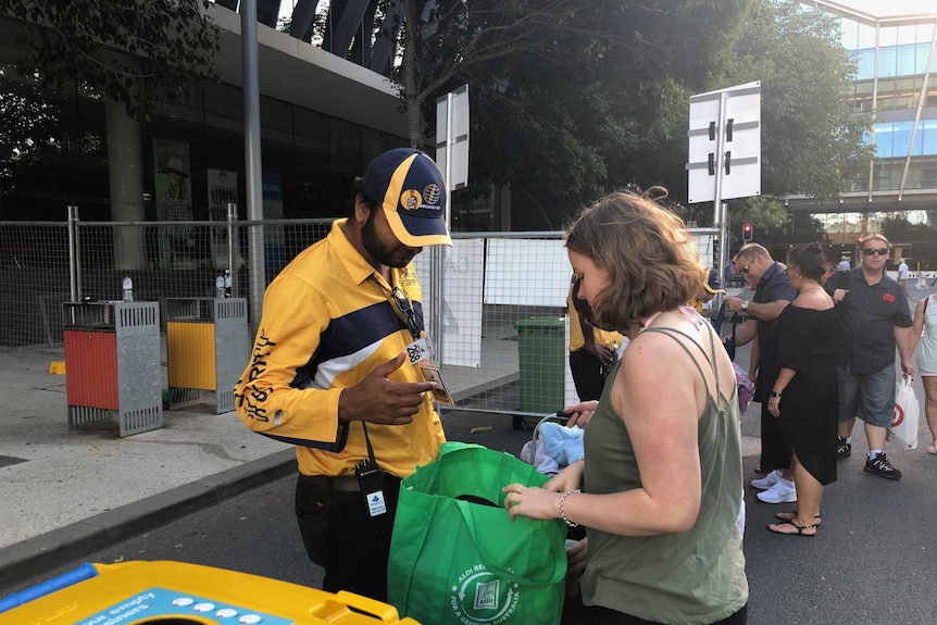 A man in flouro clothing checks a woman's green bag near a gated entry point.