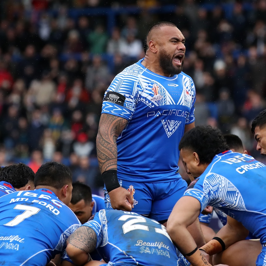 A Samoan rugby league player delivers a fierce war cry while the rest of his teammates crouch on the ground around him.  
