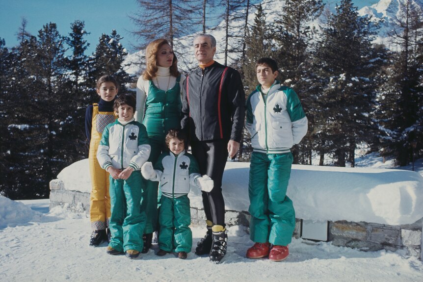A family wearing ski gear stand in a snow-covered area with pine trees behind them. It's a fairly bright day with clear skies.