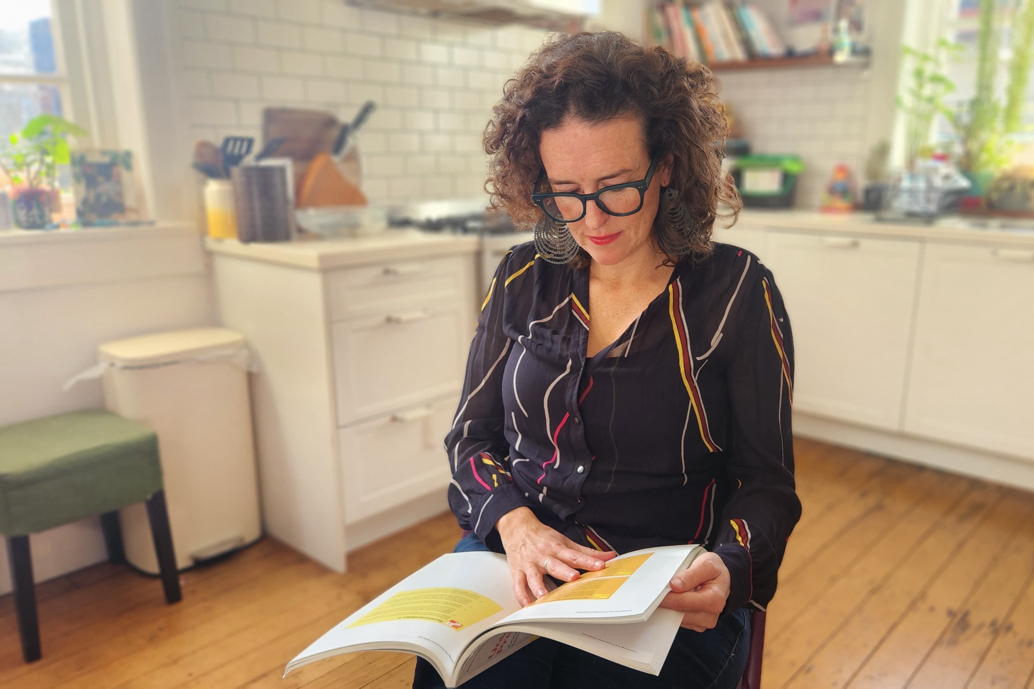 A woman with brown curly hair sites in a kitchen reading a booklet. 