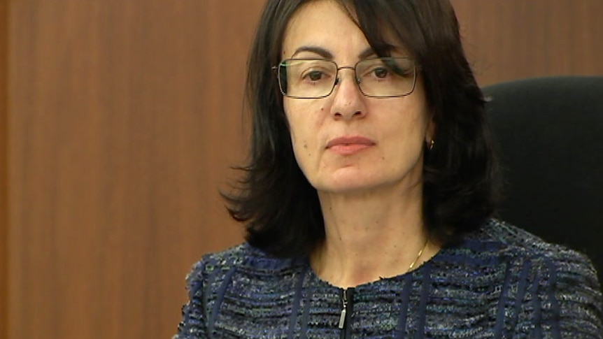 A woman in a blue top and glasses sits in court in a judge's chair.