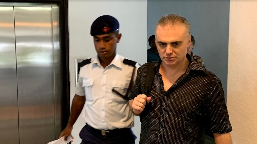 John Nikolic walks towards a court room in Suva, Fiji. A guard is beside him, Mr Nikolic carries a backpack over one shoulder.