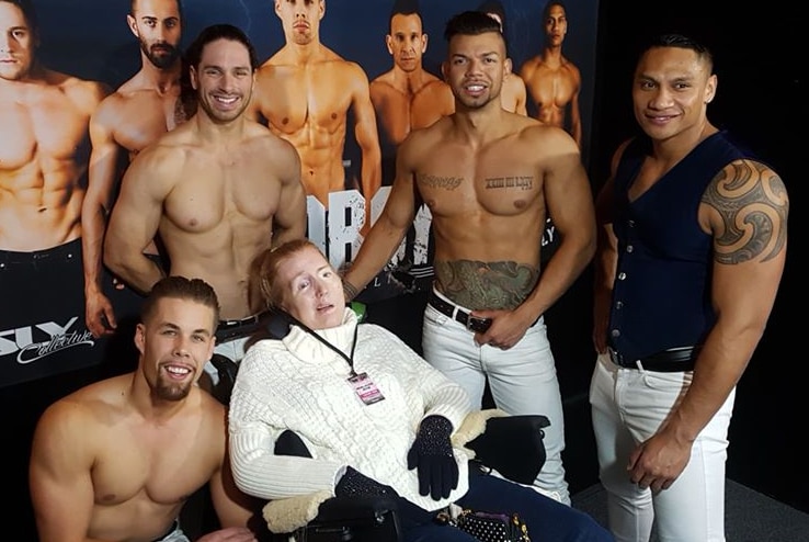 A woman in a wheelchair is surrounded by buff shirtless men smiling.